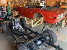 car chassis on the floor with engine and wheels, next to a body tub on a wooden platform
